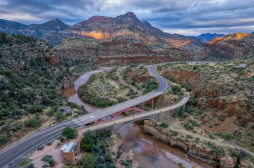 Aerial view of a winding road and bridge over a river, surrounded by rugged mountains and lush greenery.